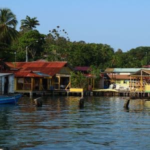 Des maisons colorées sur pilotis construites sur l'eau, avec un petit bateau bleu amarré à proximité et une forêt verte dense en arrière-plan.