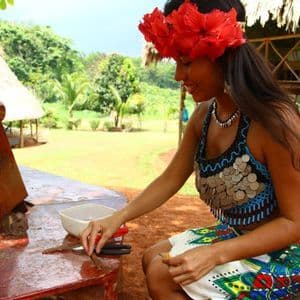 Une femme portant une coiffe de fleurs rouges et un haut perlé orné de pièces est assise à une table en extérieur, travaillant avec de petits bâtons.