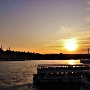 Ferries con pasajeros navegan sobre el agua al atardecer, con el perfil de la ciudad y mezquitas en silueta al fondo.
