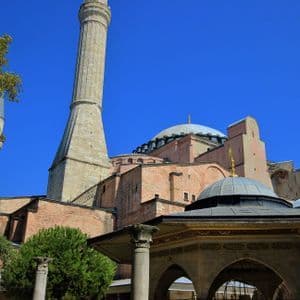 Toma en contrapicado de una mezquita histórica con grandes cúpulas y un minarete bajo un cielo azul claro, con una fuente ornamentada en primer plano.