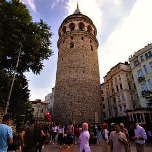 Una toma en contrapicado de una alta torre cilíndrica de piedra, rodeada de edificios, con una multitud de personas congregadas en su base.