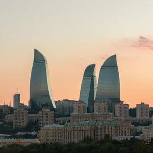 Tre moderni grattacieli di vetro a forma di fiamma svettano su uno skyline cittadino contro un cielo al tramonto dai toni caldi.