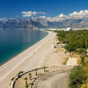 Una lunga spiaggia sabbiosa e curvilinea incontra il mare turchese, con una città costiera e un'imponente catena montuosa sullo sfondo.