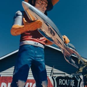 Una gran estatua de carretera de un hombre con sombrero de vaquero sosteniendo un cohete plateado frente a un letrero de la Ruta 66.