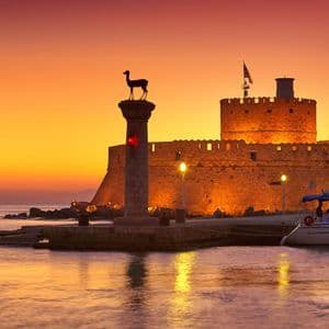 Un forte in pietra e un pilastro sormontato da una statua di cervo sono stagliati all'ingresso di un porto contro un tramonto arancione.