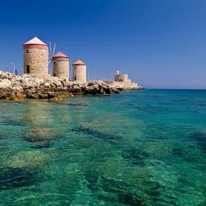 Tre mulini a vento in pietra con tetti conici rossi si ergono su una costa rocciosa accanto a un mare cristallino turchese sotto un cielo blu profondo.