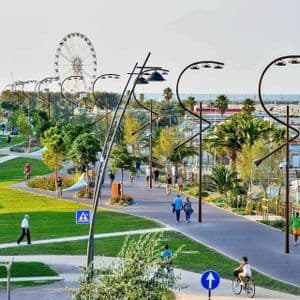 Le persone passeggiano e vanno in bicicletta lungo i sentieri di un parco costiero paesaggistico, illuminato da lampioni moderni e con una ruota panoramica in lontananza.