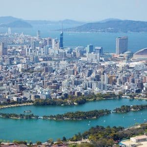 Una vista aerea di una vasta città costiera con un grande lago serpeggiante in un parco, moderni grattacieli e uno stadio a cupola sul mare.