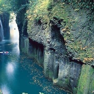 Due persone in una piccola barca remano lungo un fiume, attraversando una stretta gola con pareti rocciose muschiose e una cascata.