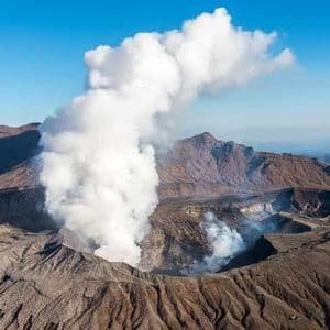 Veduta aerea di un vulcano attivo con un'ampia colonna di fumo bianco che si alza dal cratere sotto un cielo azzurro.