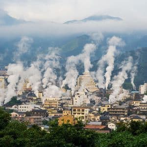 Fumi di vapore salgono dagli edifici di una città adagiata su uno sfondo di montagne verdi e nebbiose sotto un cielo nuvoloso.