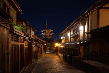 Una stretta strada in pietra di notte, affiancata da edifici tradizionali in legno, con una pagoda illuminata visibile in lontananza.