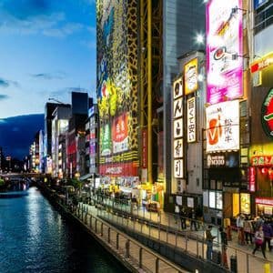 Un canale urbano al tramonto, con persone che passeggiano su un lungofiume fiancheggiato da edifici illuminati da insegne al neon.