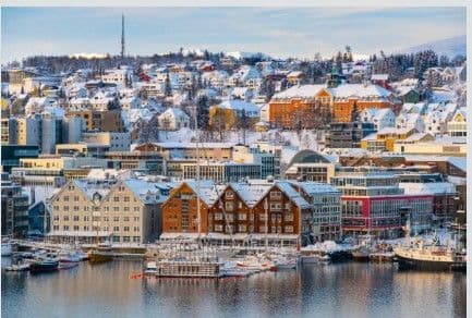 A dense, snow-covered city with colorful buildings and boats along a calm harbor during winter.