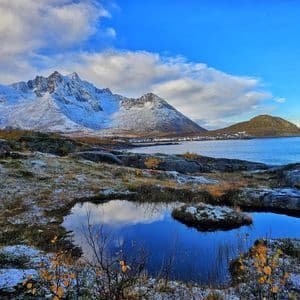 Un paisaje nevado con un estanque que refleja el cielo azul, con vistas a un pueblo costero enclavado junto a montañas cubiertas de nieve.