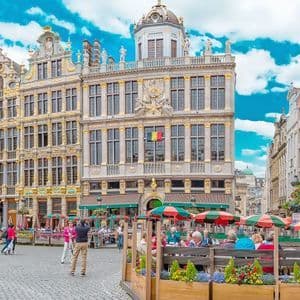 Una amplia plaza adoquinada llena de gente en cafés al aire libre, rodeada de edificios ornamentados con detalles dorados bajo un cielo azul.