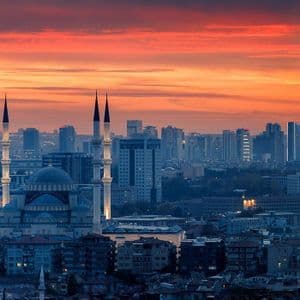 Una grande moschea con minareti illuminati si erge in un denso paesaggio urbano sotto un vibrante cielo arancione e rosso al tramonto.