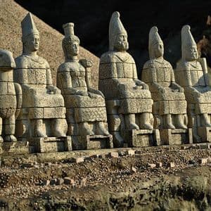 Una fila di antiche statue in pietra, corrose dalle intemperie, raffiguranti figure sedute e animali, giace su una terrazza rocciosa all'aperto.