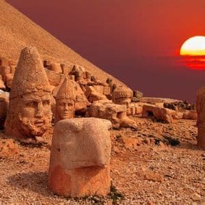 Enormi antiche teste e statue di pietra riposano su un pendio roccioso sotto un vivace tramonto rosso.