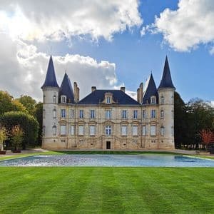 Un castillo de piedra con torretas se alza detrás de un estanque reflectante y un césped verde bajo un cielo parcialmente nublado.