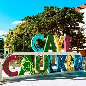 Un panneau coloré 'Caye Caulker' décoré d'illustrations d'animaux se dresse sur un sol sablonneux sous un ciel bleu clair.