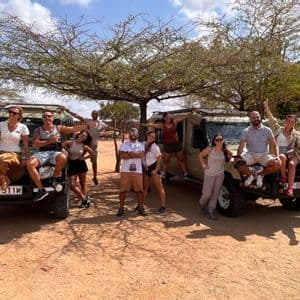 Un gruppo WeRoad in posa per una foto con due jeep safari sotto un grande albero in un paesaggio polveroso.