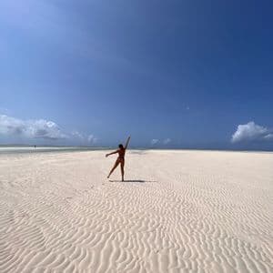 Una donna in bikini si trova su un'ampia spiaggia con sabbia bianca increspata, in posa con le braccia distese sotto un cielo azzurro.