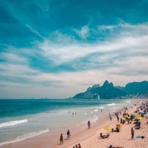 Una playa de arena concurrida con gente tomando el sol y nadando, con una ciudad y montañas de fondo bajo un cielo azul brillante.