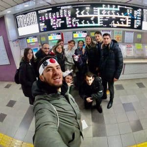A WeRoad group trip takes a group selfie in a subway station, standing in front of a large electronic map.