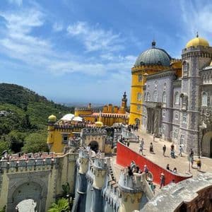 Un vivace palazzo giallo e grigio si erge su una lussureggiante collina verde, con turisti che passeggiano lungo le sue terrazze e cortili sotto un cielo azzurro.