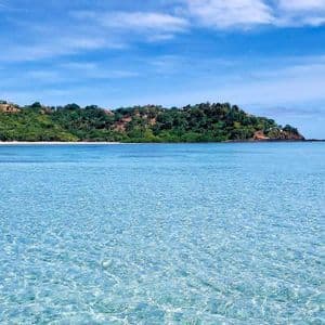Acqua cristallina color turchese luccica davanti a un'isola verde lussureggiante con una piccola spiaggia di sabbia bianca sotto un cielo blu.