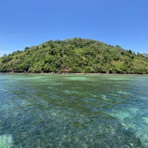 Un'isola lussureggiante e ricoperta di alberi, vista attraverso acque limpide e turchesi sotto un cielo azzurro brillante.