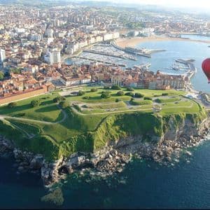 Un globo aerostático rojo flota sobre una ciudad costera con una península verde y acantilada, un puerto deportivo y una playa.