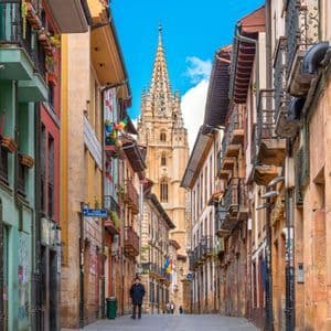 Vista de una calle estrecha con edificios coloridos, que conduce a una alta y ornamentada aguja de catedral bajo un cielo azul.