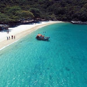 Vista aérea de una pequeña embarcación anclada en aguas turquesas cristalinas junto a una playa de arena blanca con exuberante vegetación.