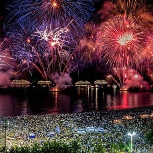 Fuegos artificiales de colores explotando sobre una playa concurrida y una bahía con varios barcos iluminados por la noche.