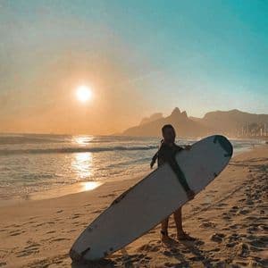Un homme en combinaison de plongée tient une planche de surf debout sur une plage de sable au coucher du soleil, avec des montagnes et une ville en arrière-plan.