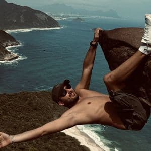Un homme torse nu, portant une casquette et des lunettes de soleil, est suspendu à une corniche rocheuse, avec en toile de fond un littoral boisé et l'océan en contrebas.