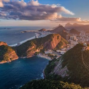Una vista aérea desde un teleférico muestra una ciudad costera anidada entre montañas verdes y el océano al atardecer.