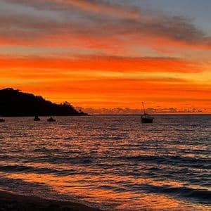 L'oceano riflette un tramonto vibrante arancione e rosso, mentre diverse barche sono stagliate contro una costa scura.