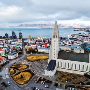 An aerial view of a large modern church overlooking a colorful coastal city with snow-capped mountains in the distance.