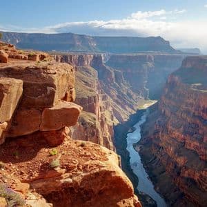 Una vista da una scogliera di roccia rossa mostra un fiume che serpeggia attraverso un canyon profondo e vasto sotto un cielo azzurro parzialmente nuvoloso.