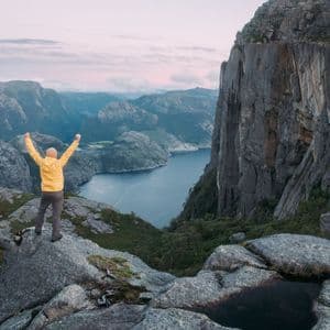 Une personne en veste jaune se tient sur un promontoire rocheux, les bras levés, surplombant un fjord et un paysage montagneux.