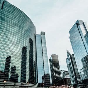 Vista dal basso di imponenti grattacieli di vetro, con una facciata curva che riflette altri edifici contro un cielo coperto.