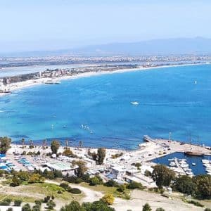 Una vista aérea de un paisaje costero con una playa de arena blanca curvada, un puerto deportivo y una bahía azul brillante con veleros.