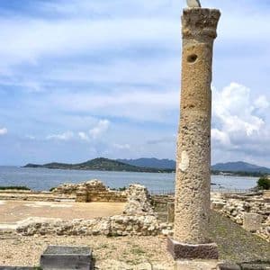 Una gaviota se posa en una columna de piedra erosionada en un sitio arqueológico con vistas al mar bajo un cielo parcialmente nublado.