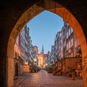 Eine gepflasterte Straße, gesäumt von historischen Gebäuden, unter einem warmen Backsteinbogen in der Dämmerung gesehen.