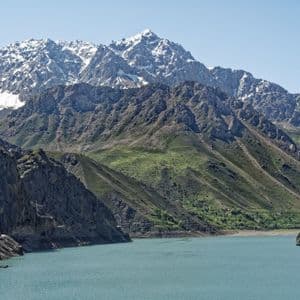 Un lago turchese in una valle circondata da ripidi pendii verdi e montagne rocciose, con cime innevate sullo sfondo.