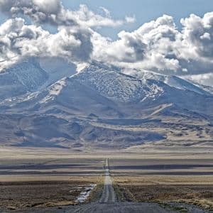 Una lunga e dritta strada sterrata si estende attraverso una vasta pianura arida verso una catena montuosa innevata sotto un cielo nuvoloso.