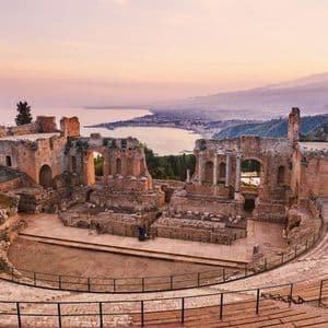 Un antico anfiteatro in pietra in rovina, che si affaccia su una costa e una grande montagna in lontananza al tramonto.